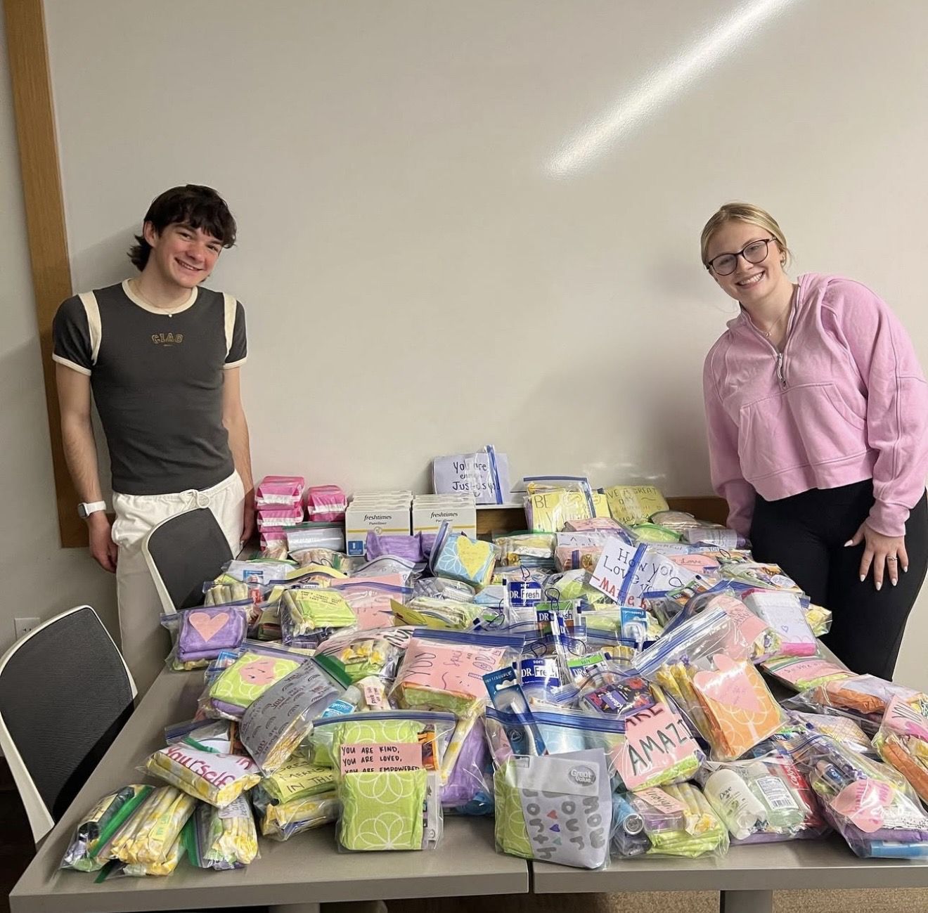 Two empowHER members smiling beside a table covered in packed hygiene kits with handwritten notes